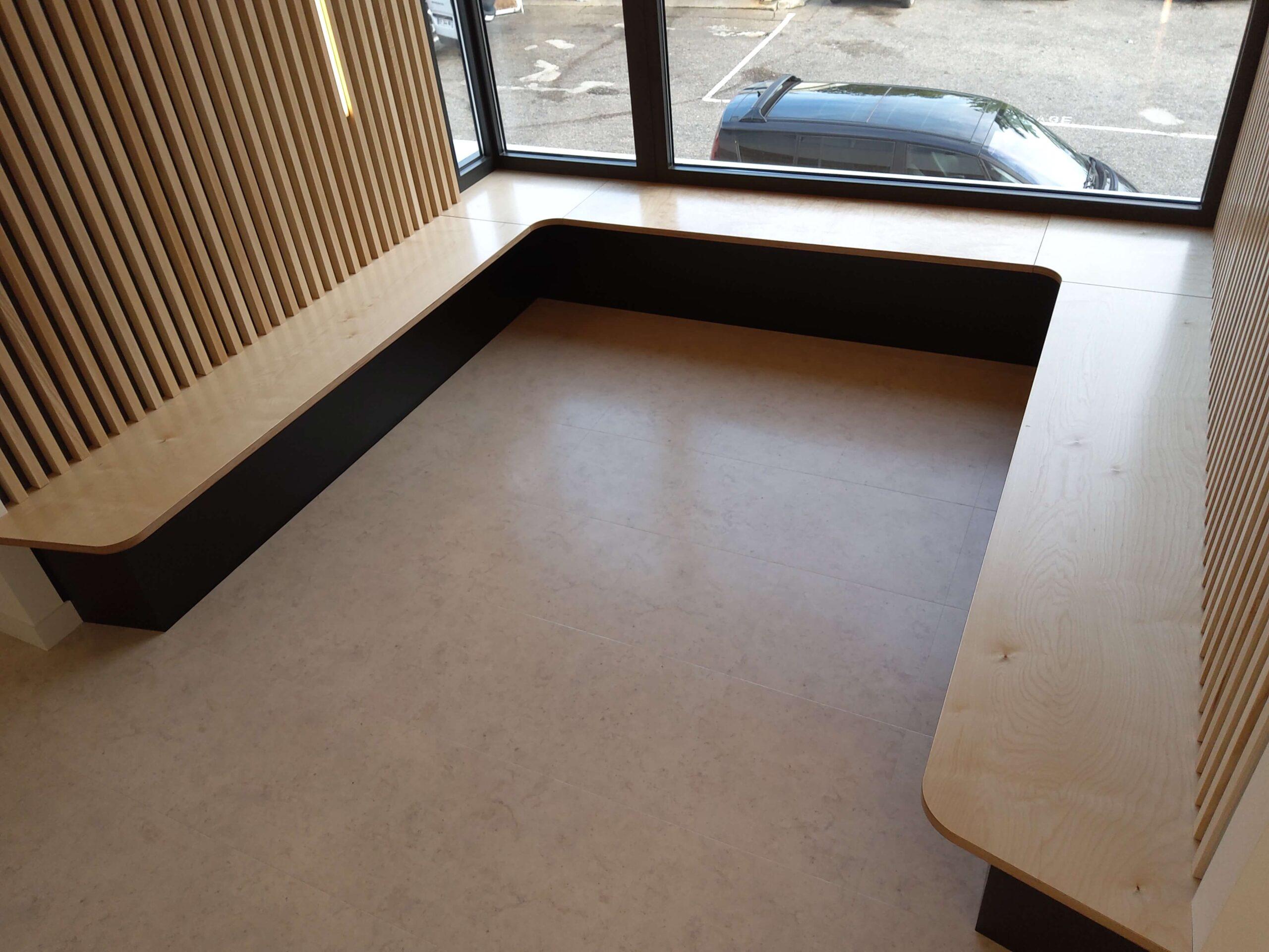 Espace-d'attente-centre-médical Menuiserie en bois de l'espace d’attente avec des bancs en contre-plaqué bouleau vernis, soubassement en stratifié noir, panneaux de dossier et de plafonds en tasseaux frêne vernis naturel avec fixation invisible sur panneaux mélaminé finition touch et intégration de bandeaux Led en coordination avec le lot électricité.
