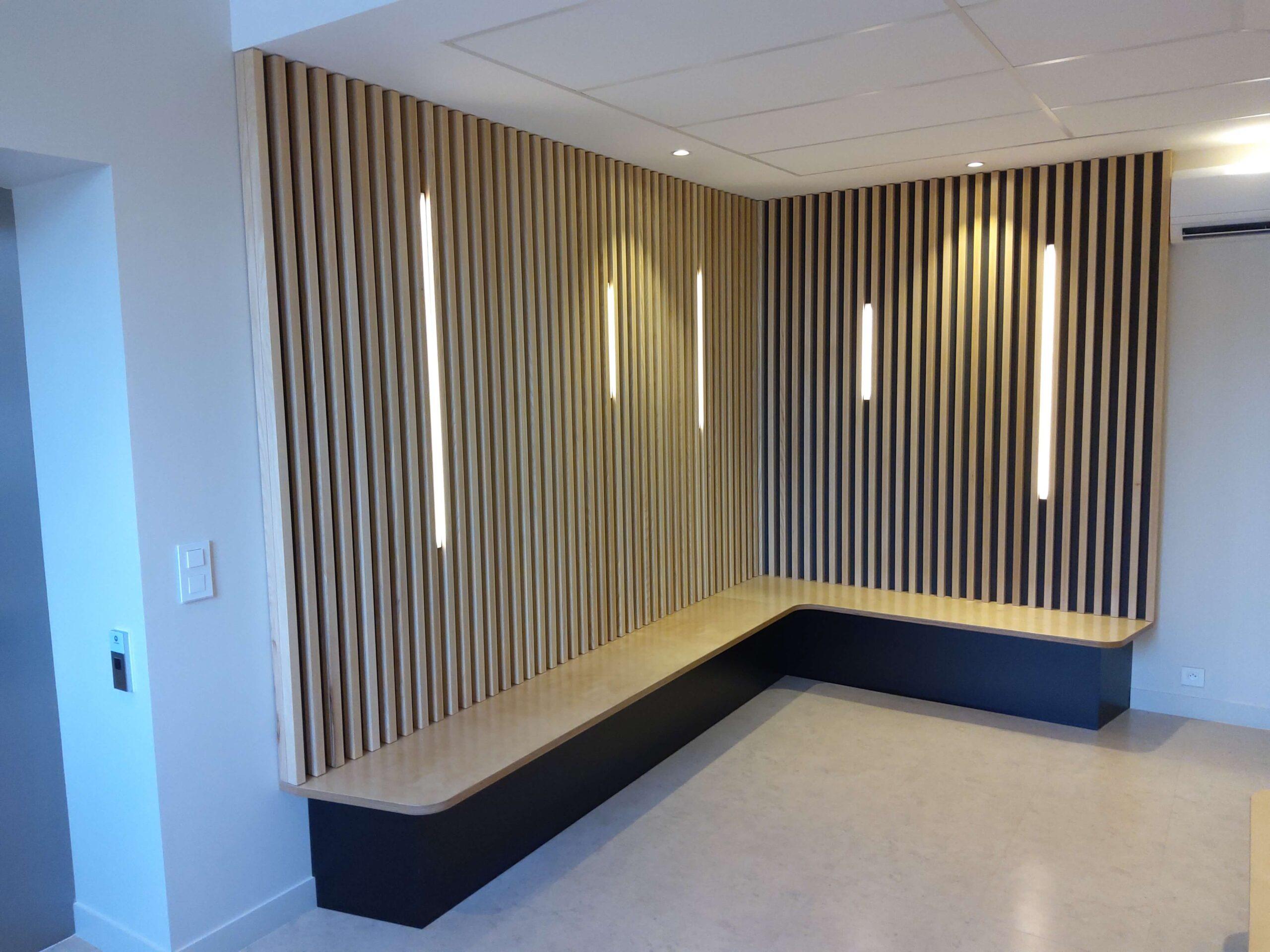 salle-d'attente-médical Menuiserie en bois de la salle d’attente avec des bancs en contre-plaqué bouleau vernis, soubassement en stratifié noir, panneaux de dossier et de plafonds en tasseaux frêne vernis naturel avec fixation invisible sur panneaux mélaminé finition touch et intégration de bandeaux Led en coordination avec le lot électricité.